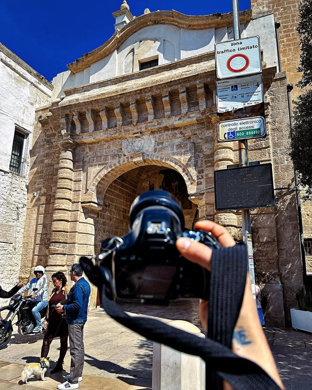 A person holding a camera up in front of an old stone archway with several people walking nearby, a woman with a dog, and a person on a scooter. There are street signs in Italian indicating a limited traffic zone and accessible electronic control.