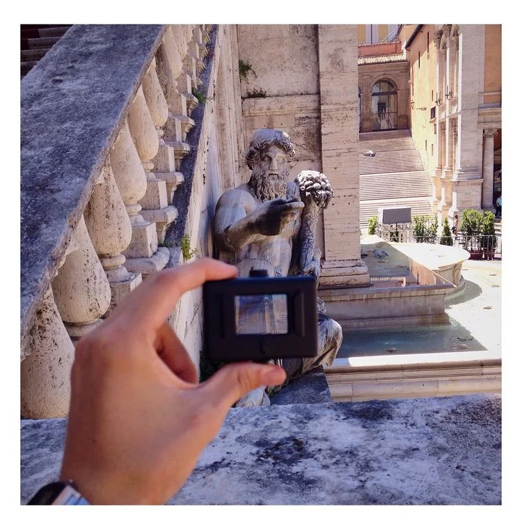 Person taking a picture of a stone sculpture of a bearded man holding a bunch of grapes, situated near classical architecture with stairs and a fountain in the background.