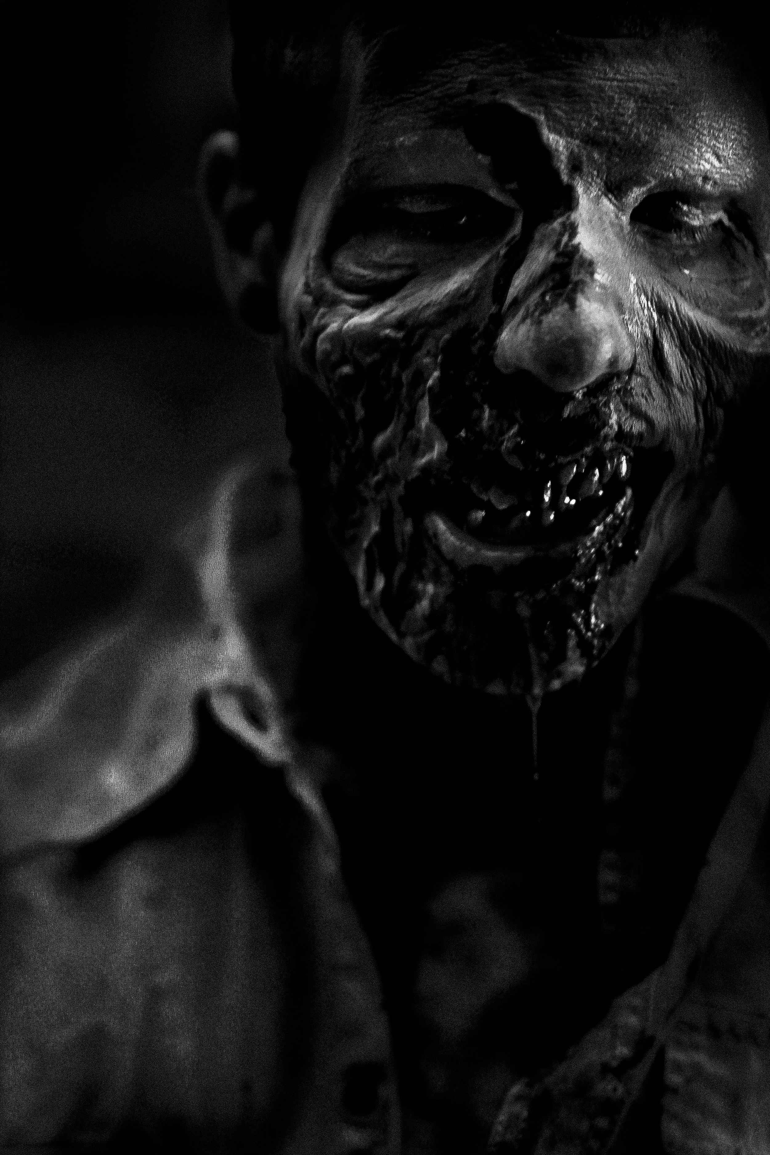 A person with face paint resembling a skull, showing teeth and dark eyes in a close-up shot, black and white. 