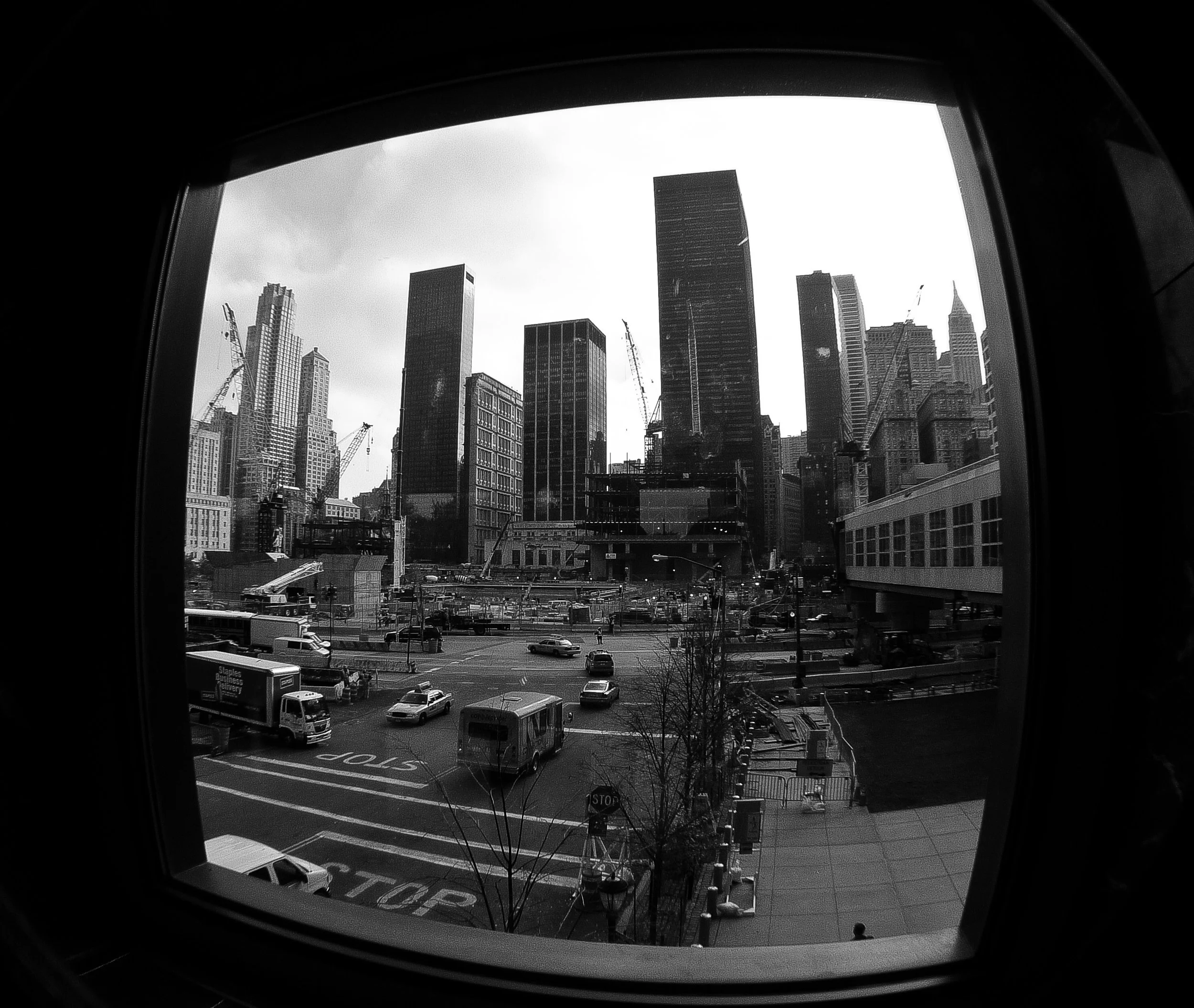 Black and white photo of a city skyline seen through a circular window, with buildings, cranes, and parked cars in the foreground.
