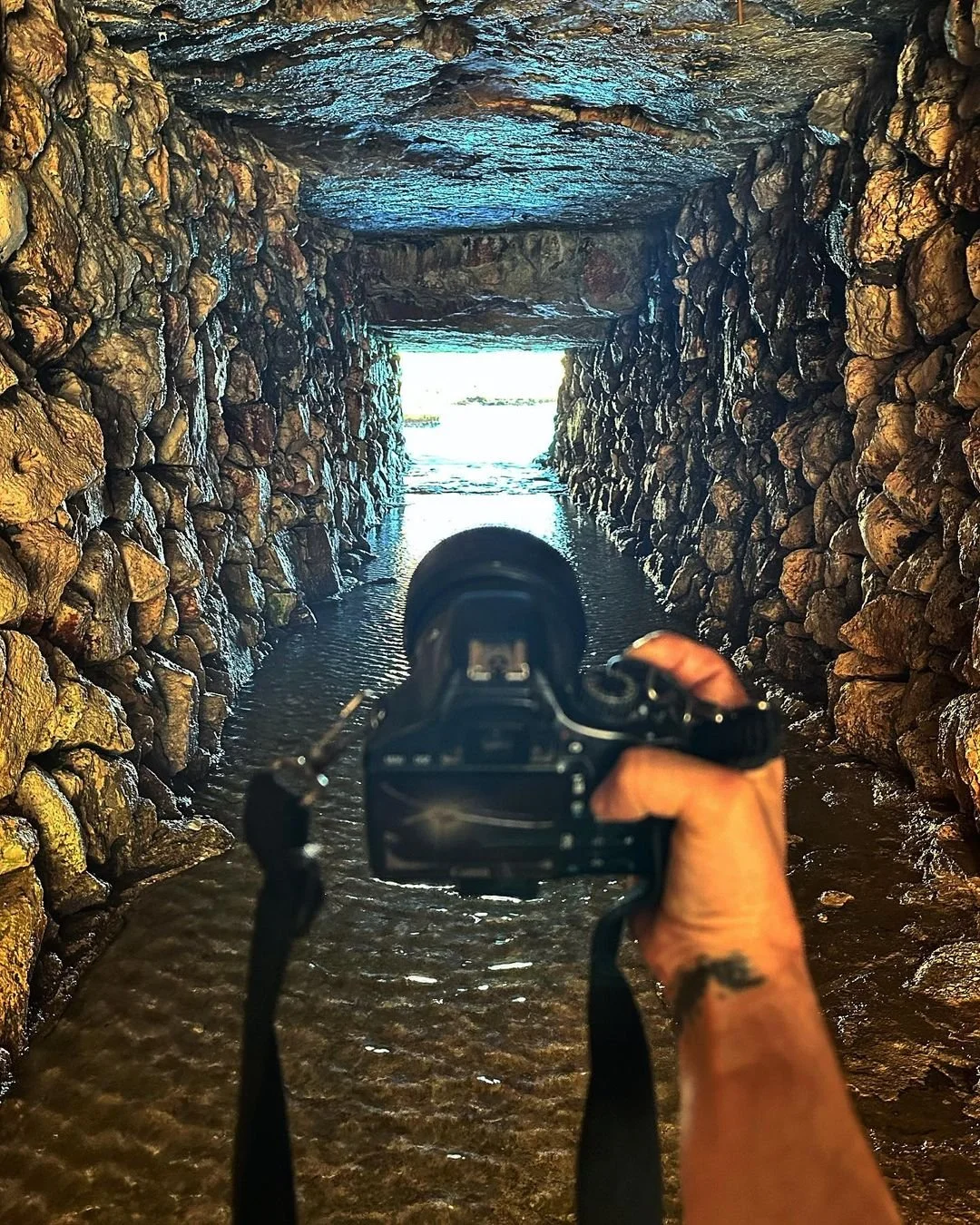 A person holding a camera captures a view of a stone tunnel or bridge with water flowing underneath, leading to an opening with bright light outside.