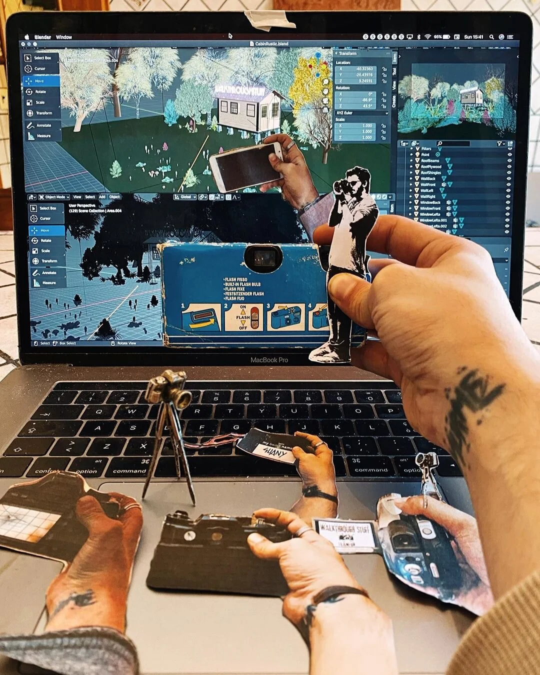 A person holding a cutout figure of a woman taking a photograph in front of a laptop with a digital environment on the screen. The scene includes multiple hands, small cameras, and photography-related objects placed on the laptop. The digital environ