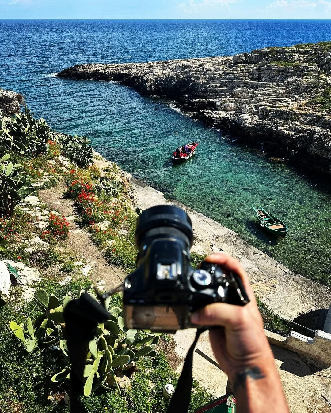 A person holding a camera pointed towards a rocky cove with two boats floating in clear, turquoise water, and a blue ocean extending to the horizon in the background. There are cacti and plants on the rocky terrain in the foreground.