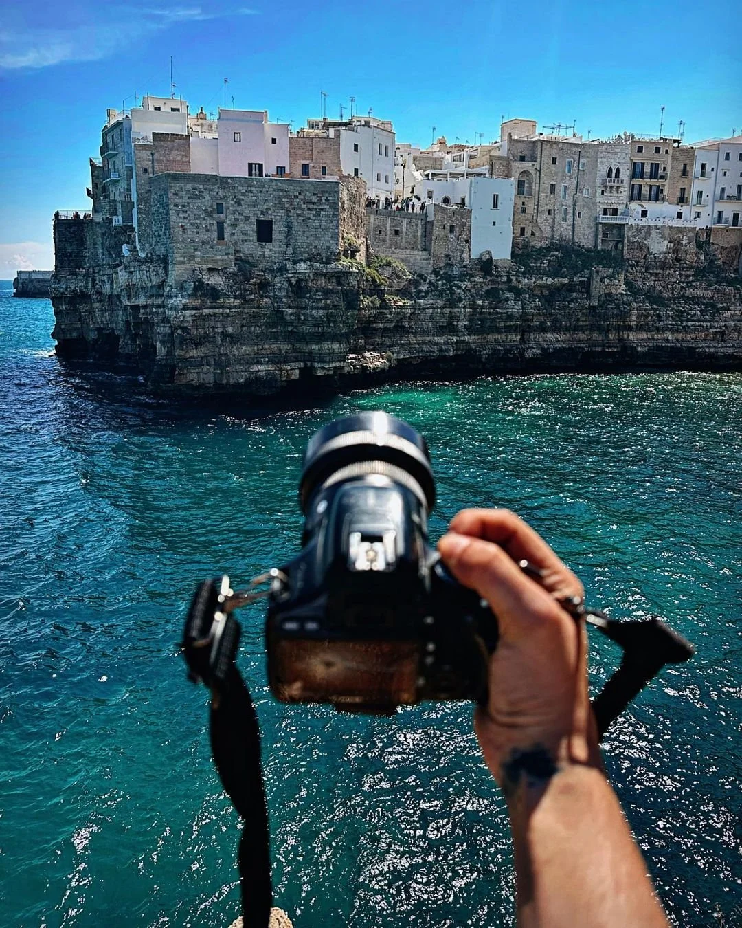 A person holding a camera taking a photo of a coastal town with white buildings and rocky cliffs over turquoise water.