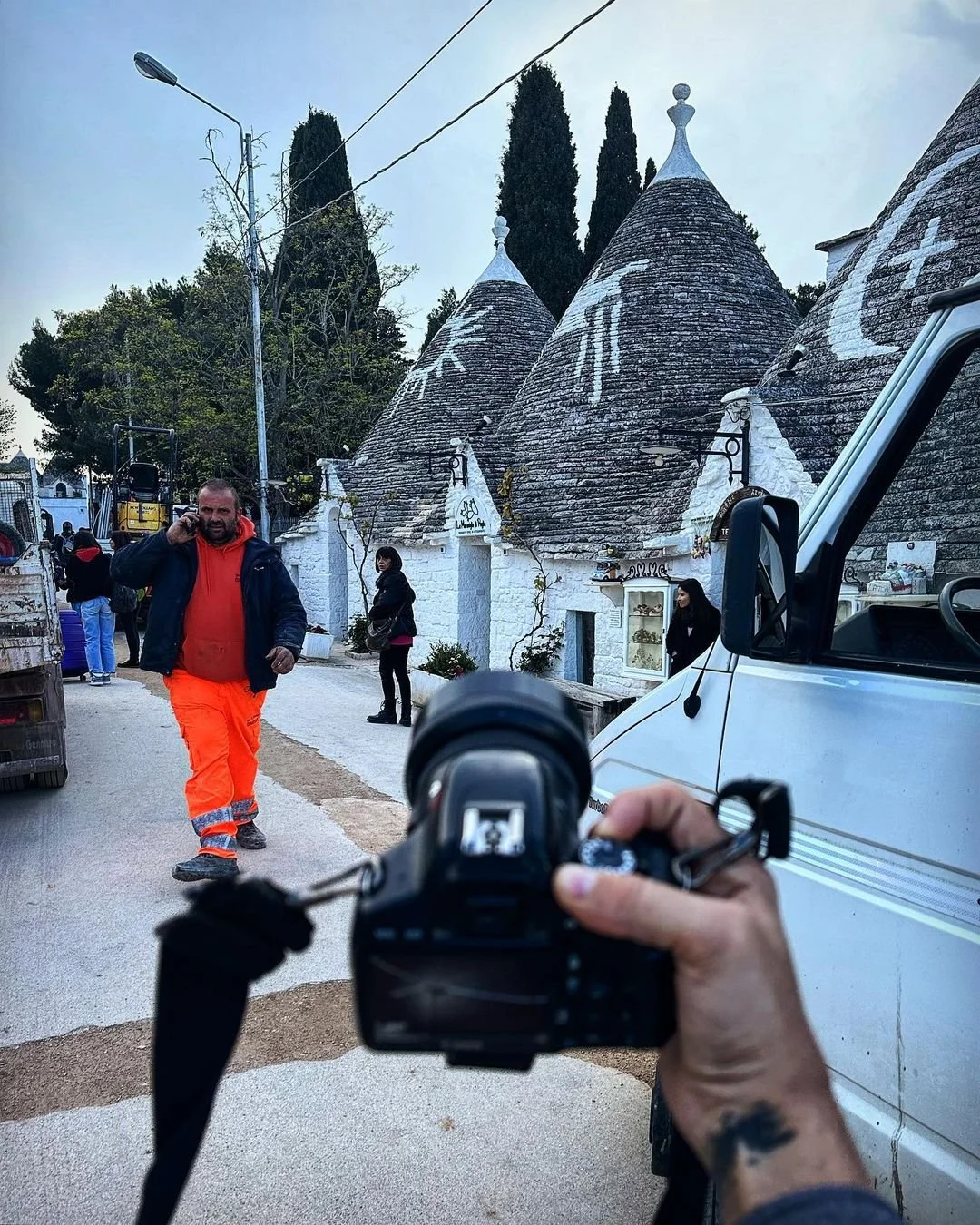 Photographer taking a picture of a man in bright orange pants and a red hoodie walking on a street with traditional white cone-shaped buildings in the background.