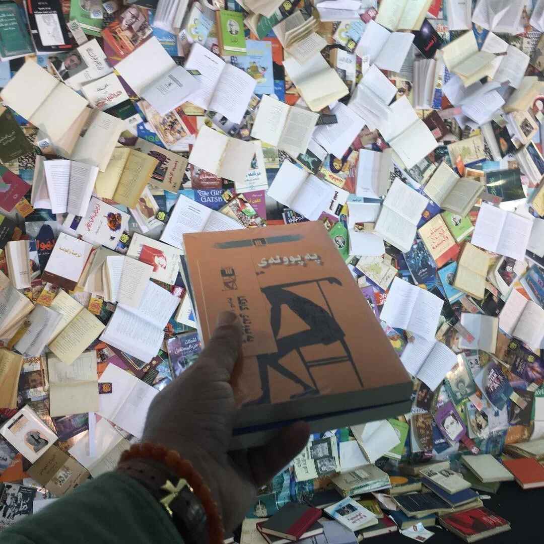 A person holding an orange book with silhouette of a chair on the cover, in a space filled with many open books and scattered pages.