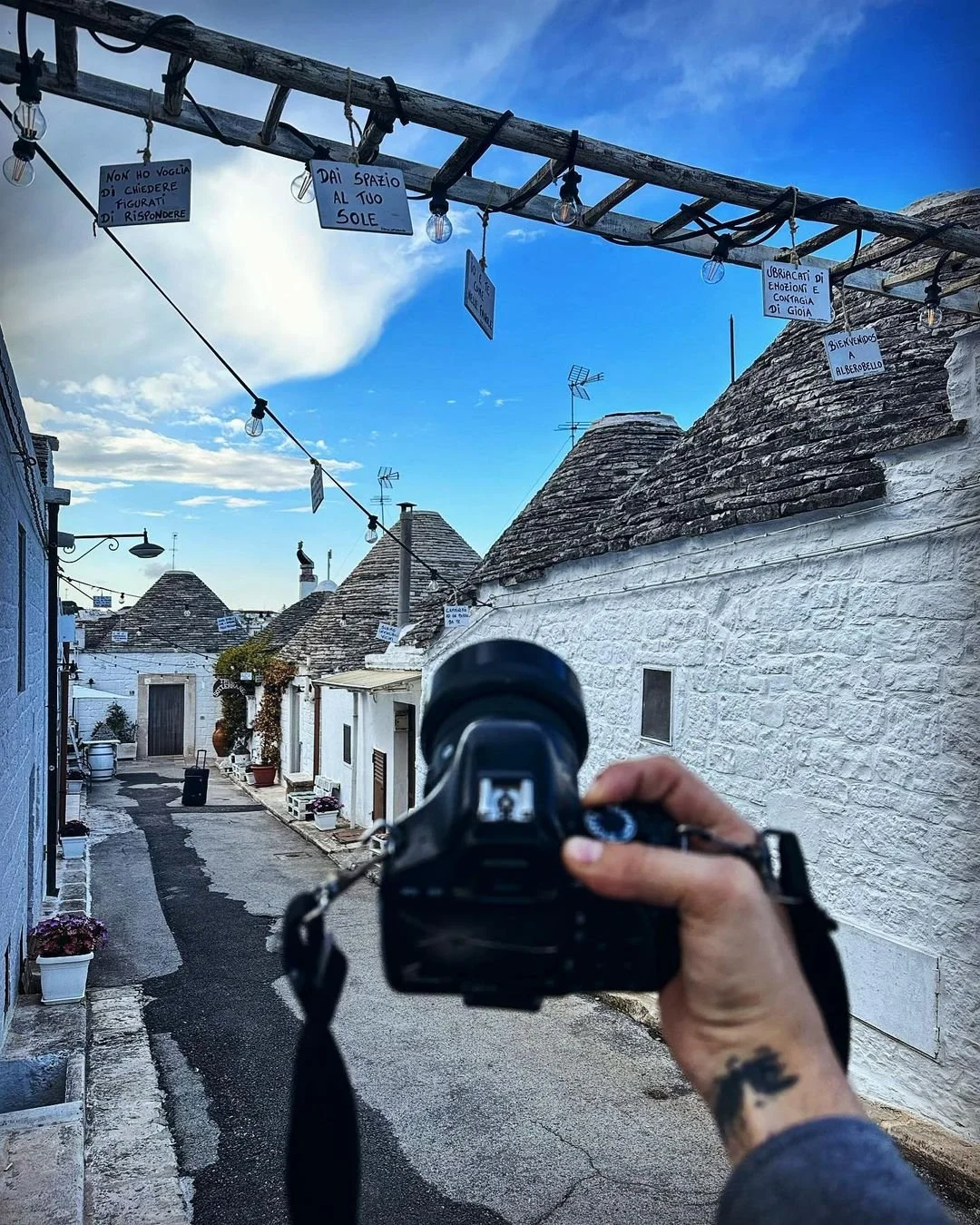 A narrow street in a village with white stone buildings and cone-shaped roofs. A camera is being held by a person in the foreground, taking a photo of the scene. String lights and handwritten signs are hanging above the street, with one sign reading 'Dai spazio al tuo sole'.