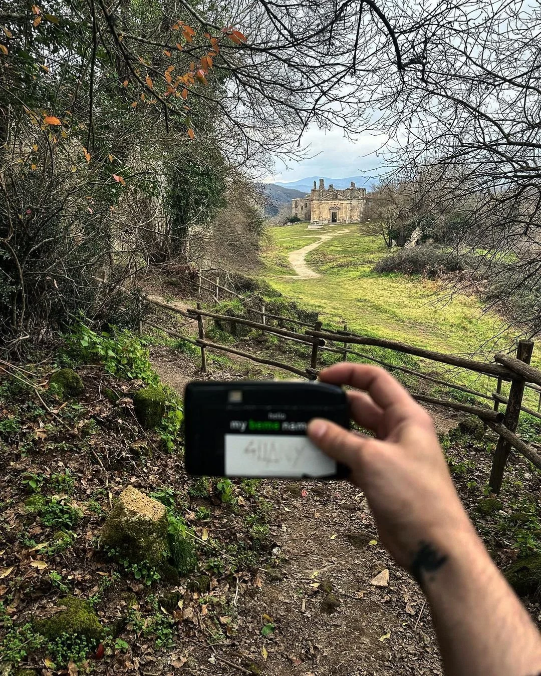 Person holding a police badge in front of a trail leading to an ancient stone building surrounded by trees.