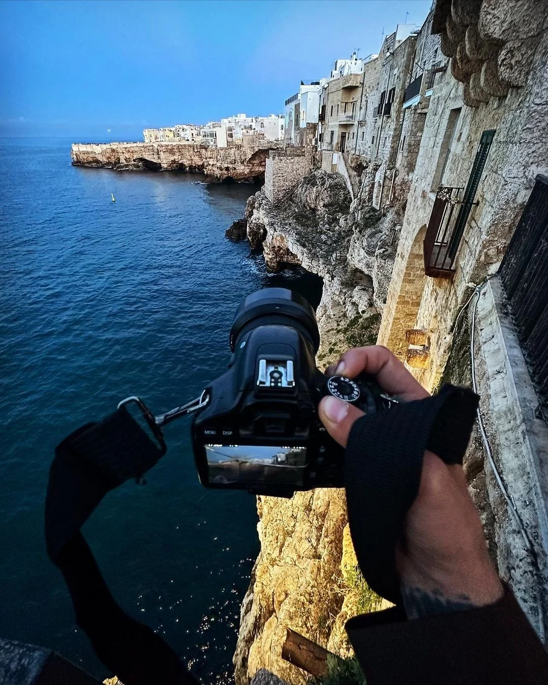 Person holding a camera taking a picture of a coastal cliffside with buildings on top, overlooking the ocean under a clear blue sky.