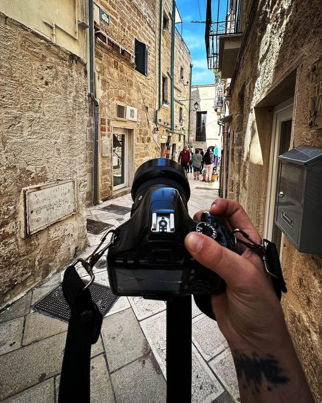 A photographer's point of view capturing a narrow street with stone buildings, showing a camera held by a person's hand in the foreground and a group of people walking ahead under a bright blue sky.