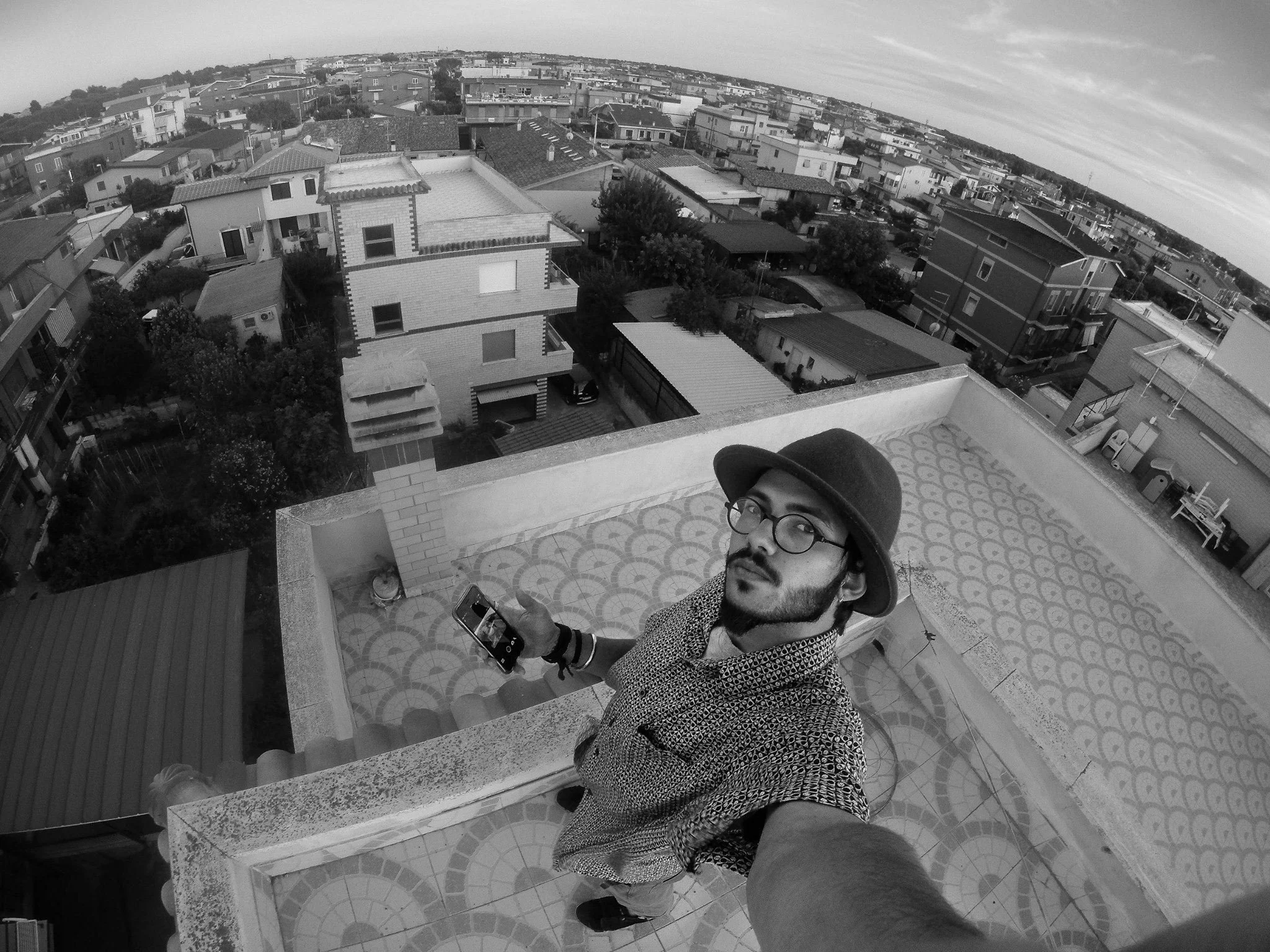 A man in a patterned coat and glasses, holding a phone, standing on a balcony with a patterned tiled floor, overlooking a cityscape of houses and buildings, under a partly cloudy sky.