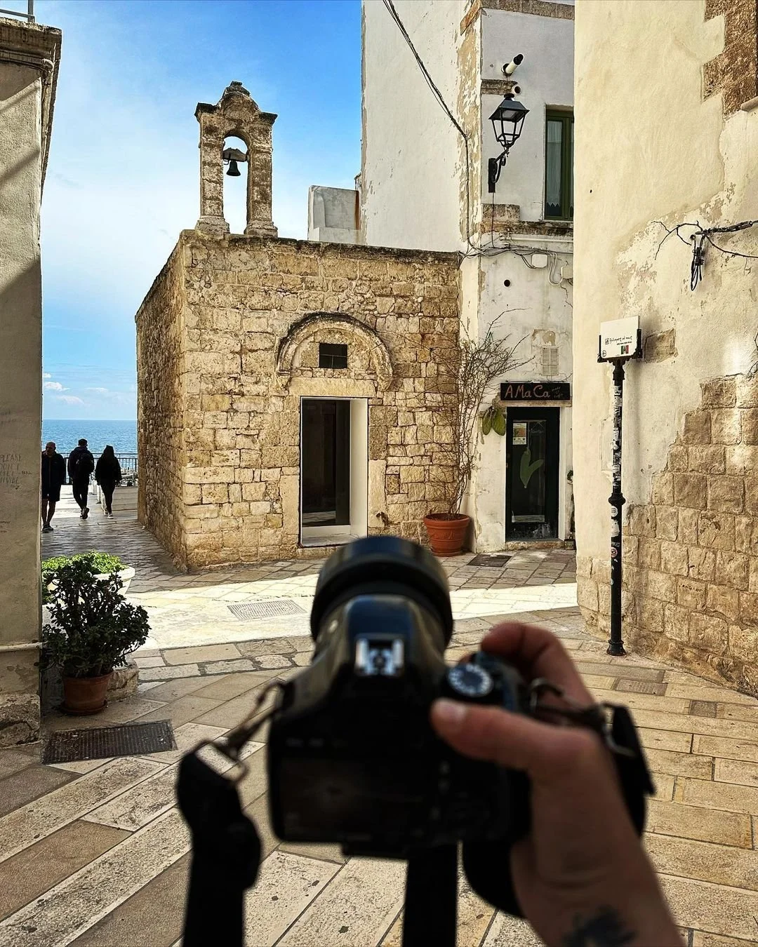 A person holding a camera in front of a small, stone building with a bell tower on top, situated in a European-style street with white and beige buildings, potted plants, and a view of the sea in the background.