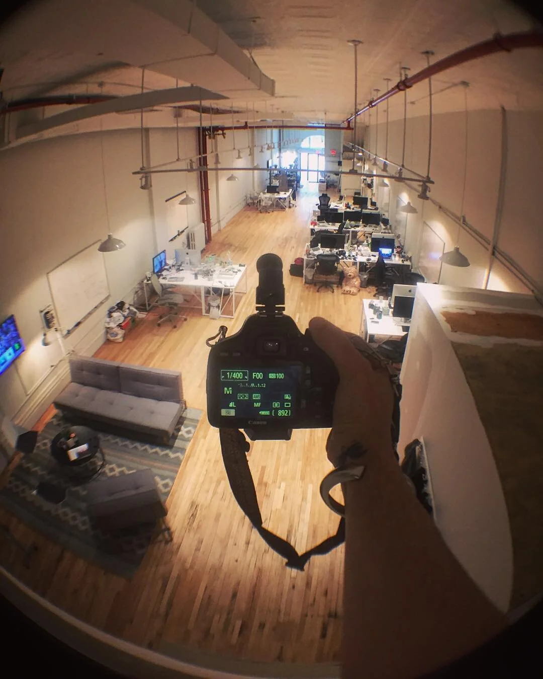 Looking down from a high vantage point, a person holds a camera capturing a spacious open office with desks, computers, and a living room area with couches and a television.