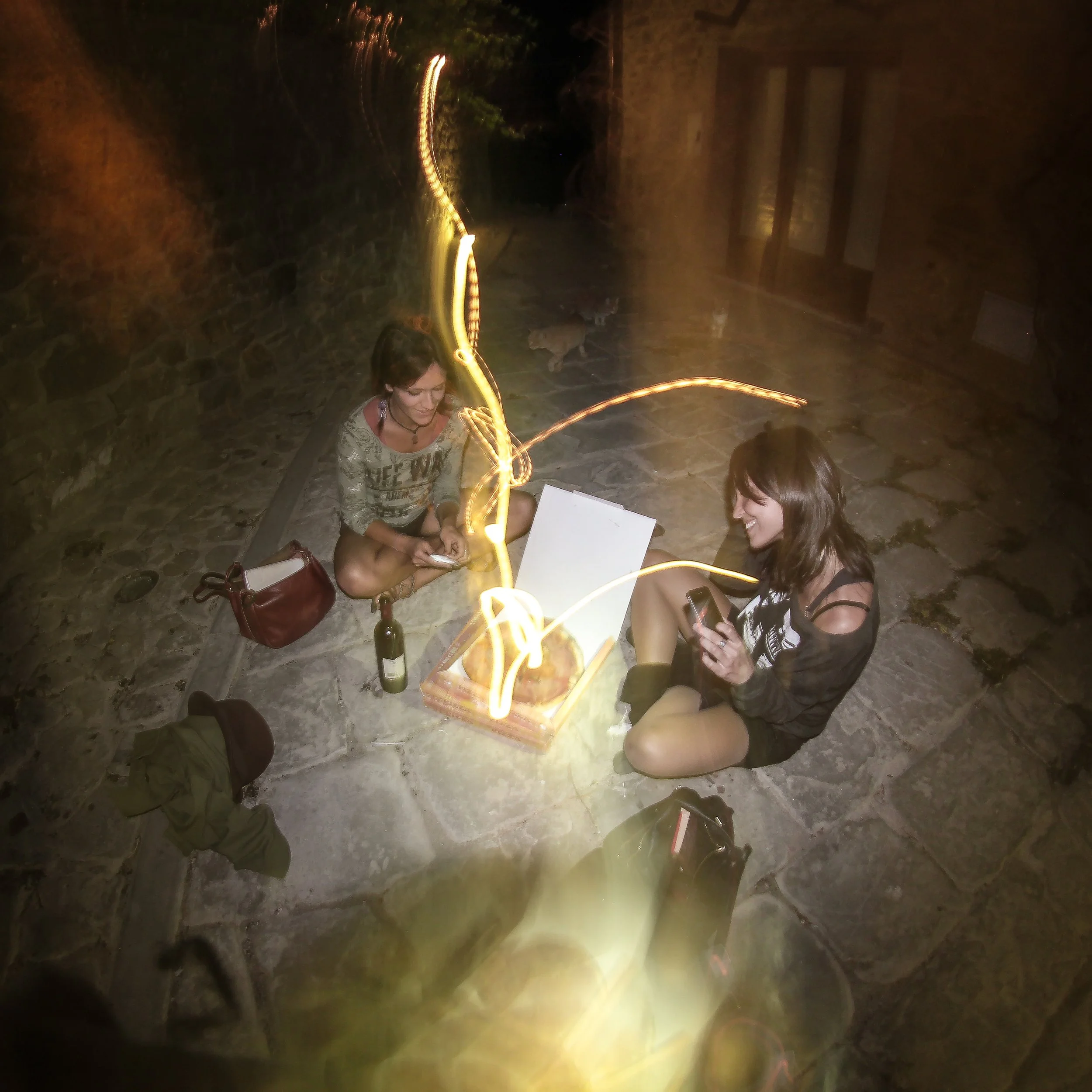 People sitting around a table with a bottle of wine, some glasses, and a book, illuminated by string lights at night.
