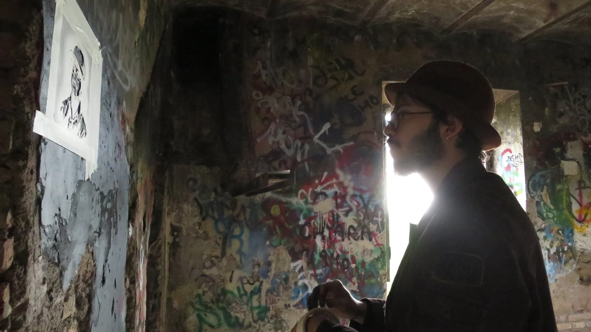 A man wearing glasses, a hat, and a dark jacket stands sideways inside a graffiti-covered underground tunnel with a rough stone wall. Bright light shines from behind him, illuminating the area and casting his shadow.