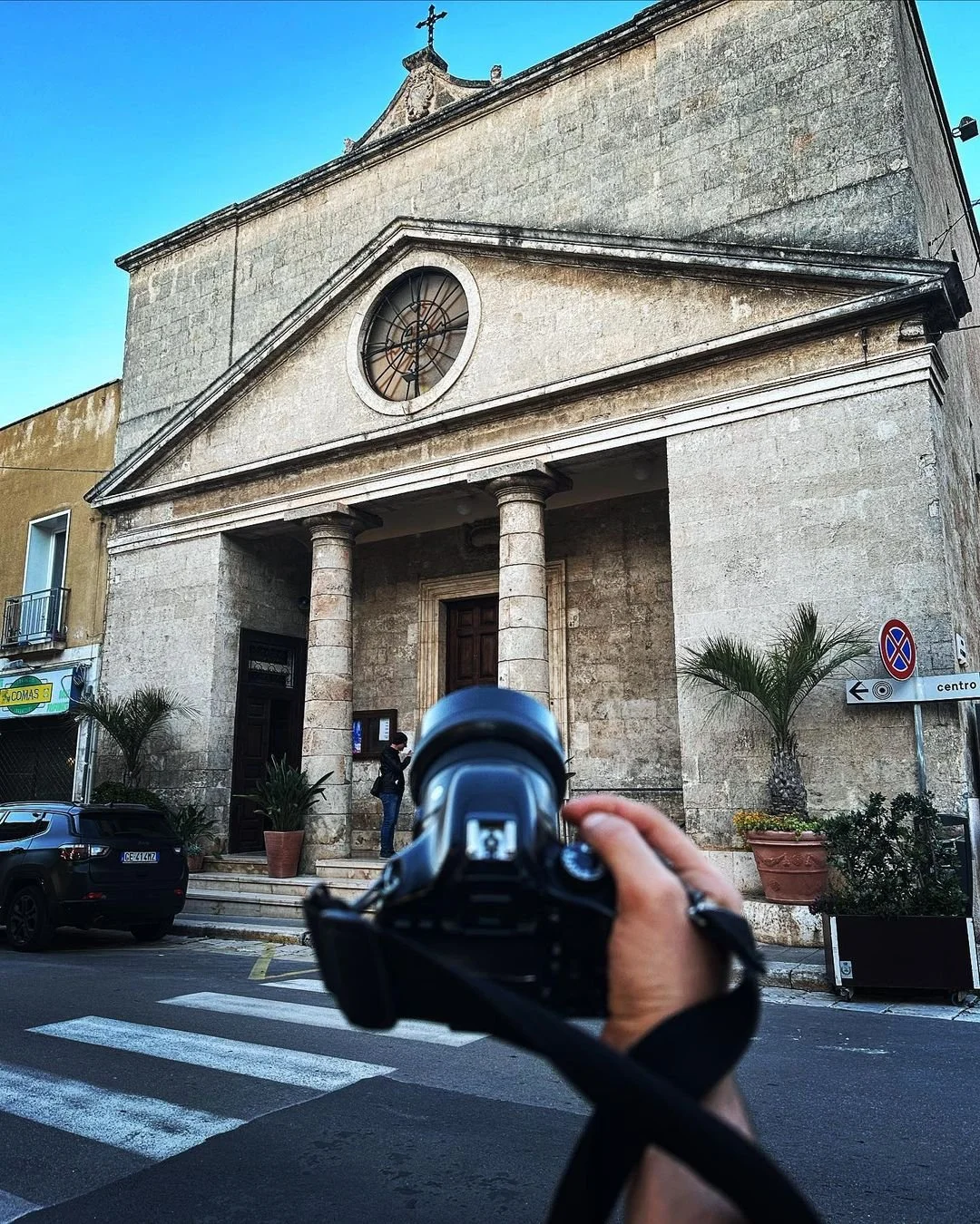 A person holding a camera is taking a photo of a stone church with columns and a clock on its facade. The church has a cross on the top and two potted plants are near the entrance. There is a street with parked cars and a no parking sign to the right.