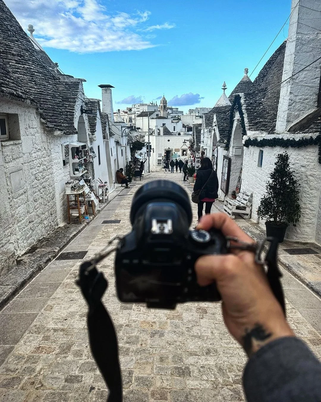 Photo of a narrow street with white stone buildings, some with shops, and people walking and sitting. The person taking the picture is holding a camera, with the back of their hand and a tattoo visible. The sky is partly cloudy and the scene appears to be in a historic town.