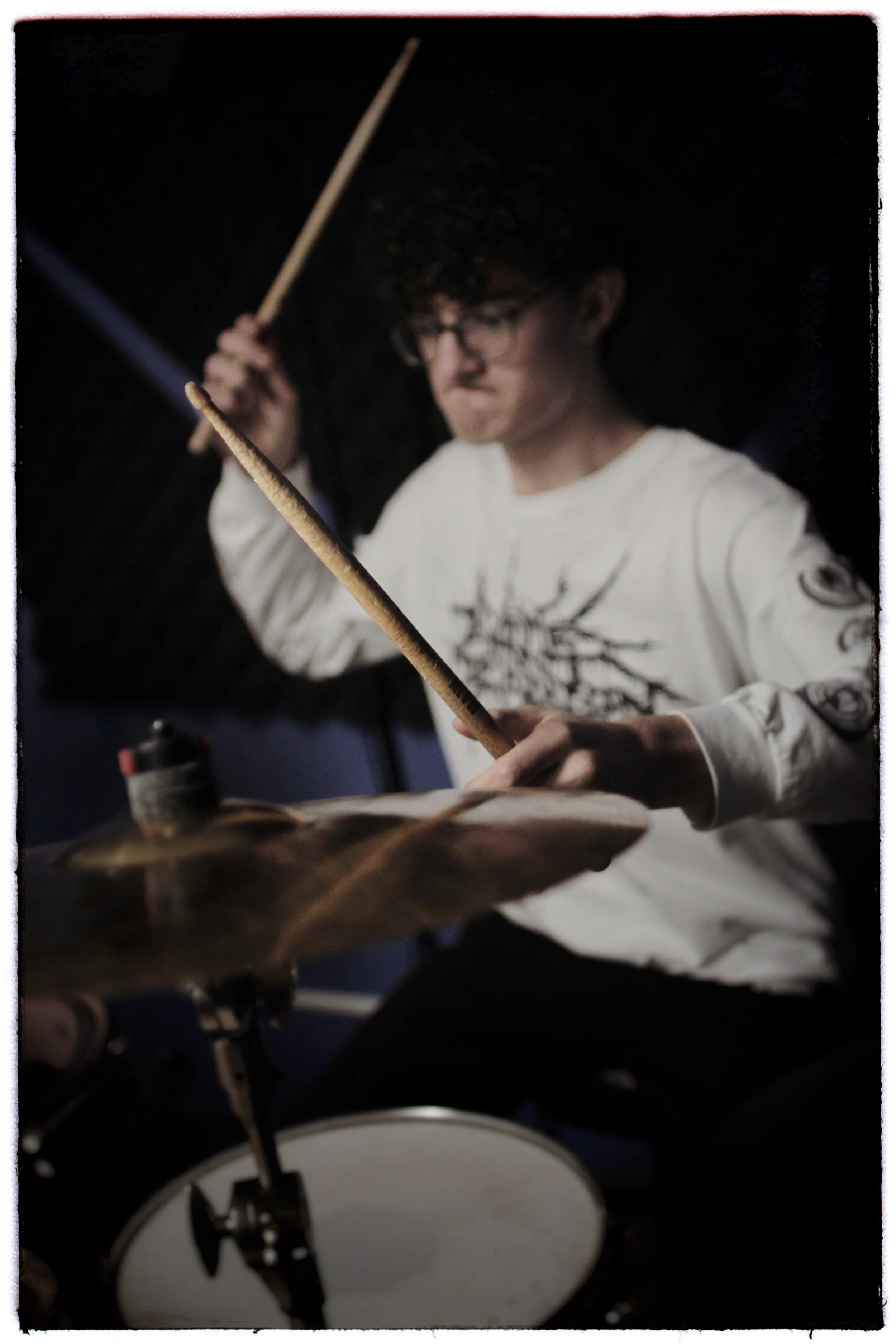 A person with glasses playing drums, holding drumsticks, in a dark setting.