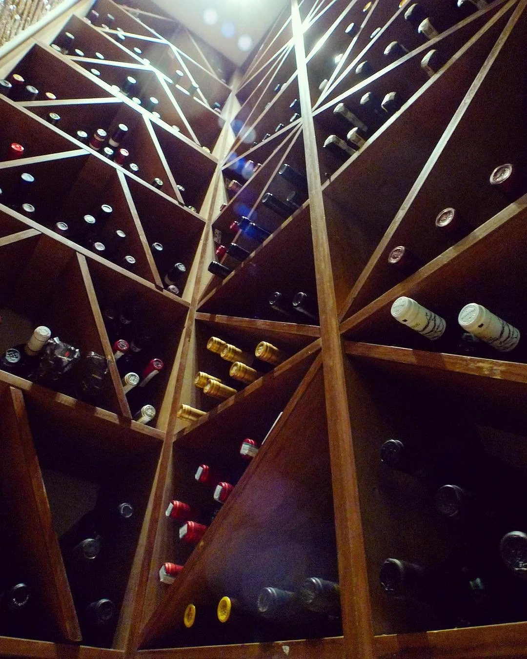 Wine bottles stored horizontally in a wooden wine rack with multiple compartments, viewed from below.