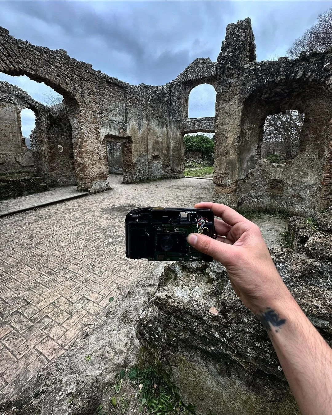 A person holding a camera, taking a photo of the ruins of an old stone building with arched windows and doorway, under a cloudy sky.