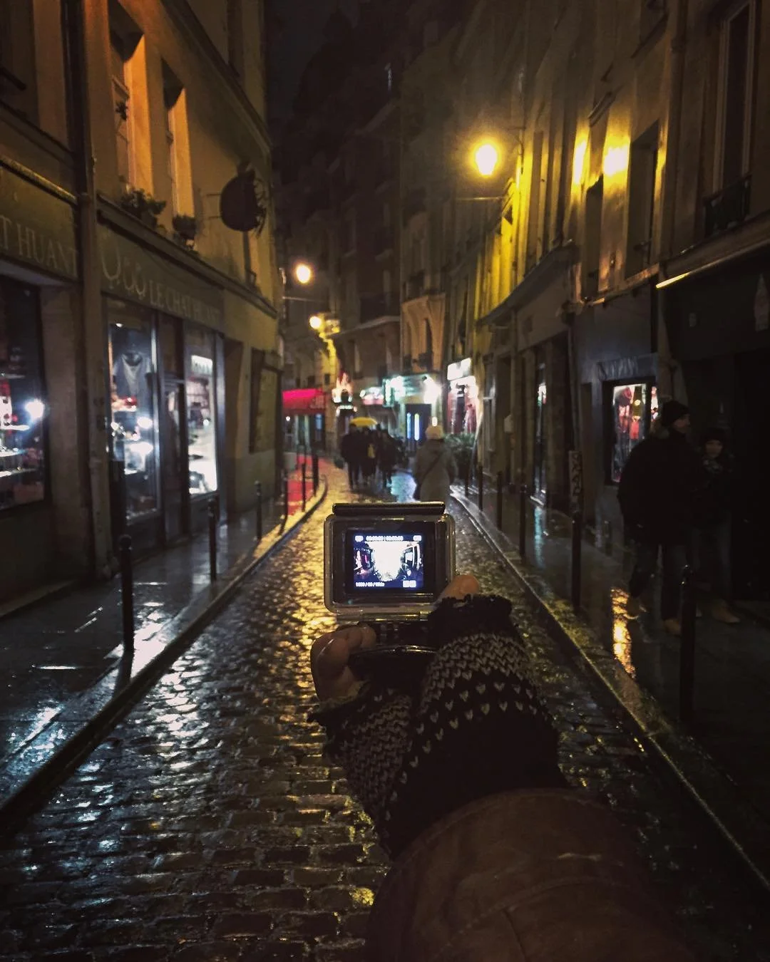 Nighttime street scene with wet cobblestone pavement, illuminated shopfronts, and pedestrians with umbrellas. A person is taking a photo with a camera, capturing the scene.
