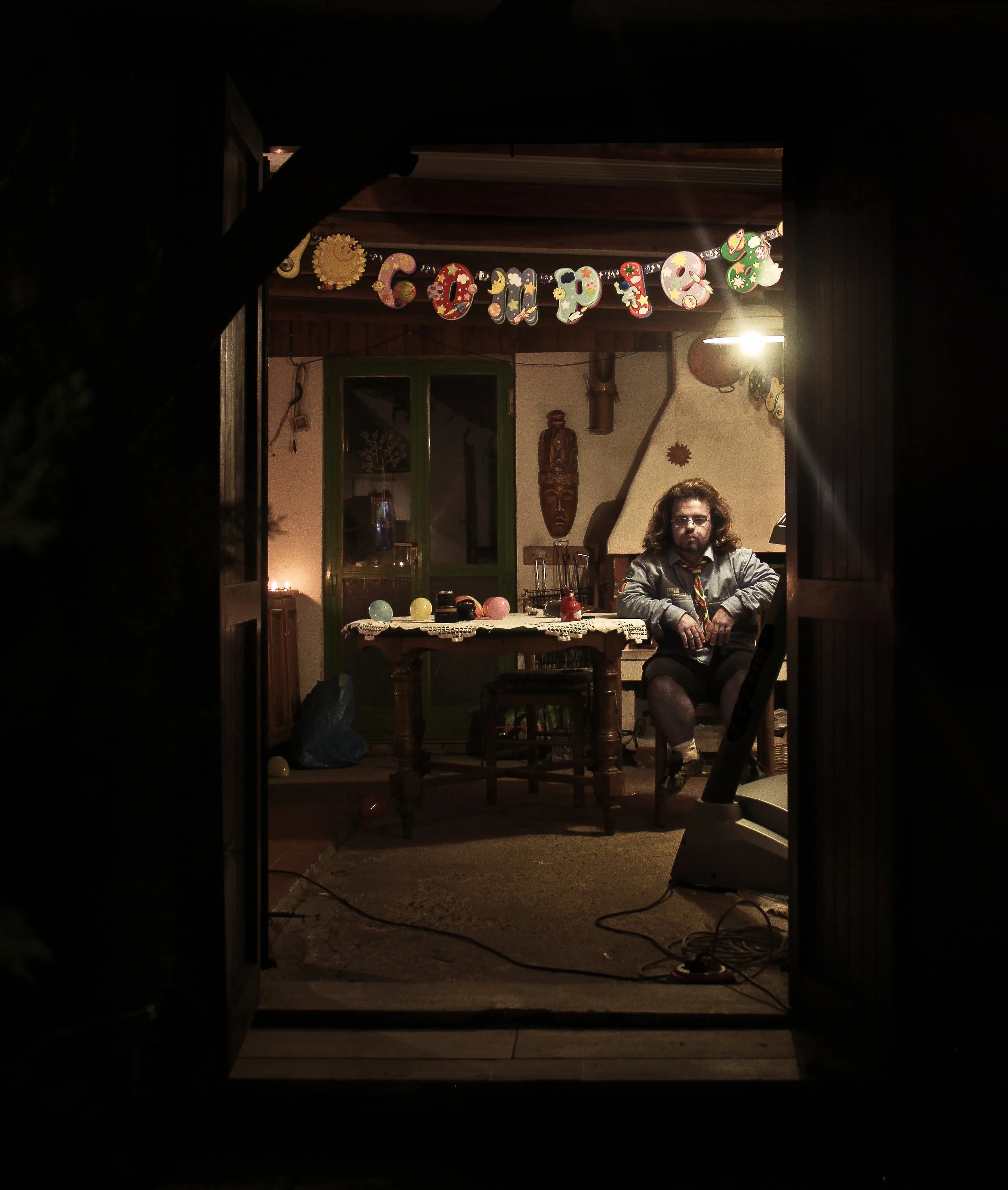 A person sitting at a wooden dining table in a cozy, decorated room, with birthday decorations including balloons and a "Congratulations" banner, seen through a doorway at night.