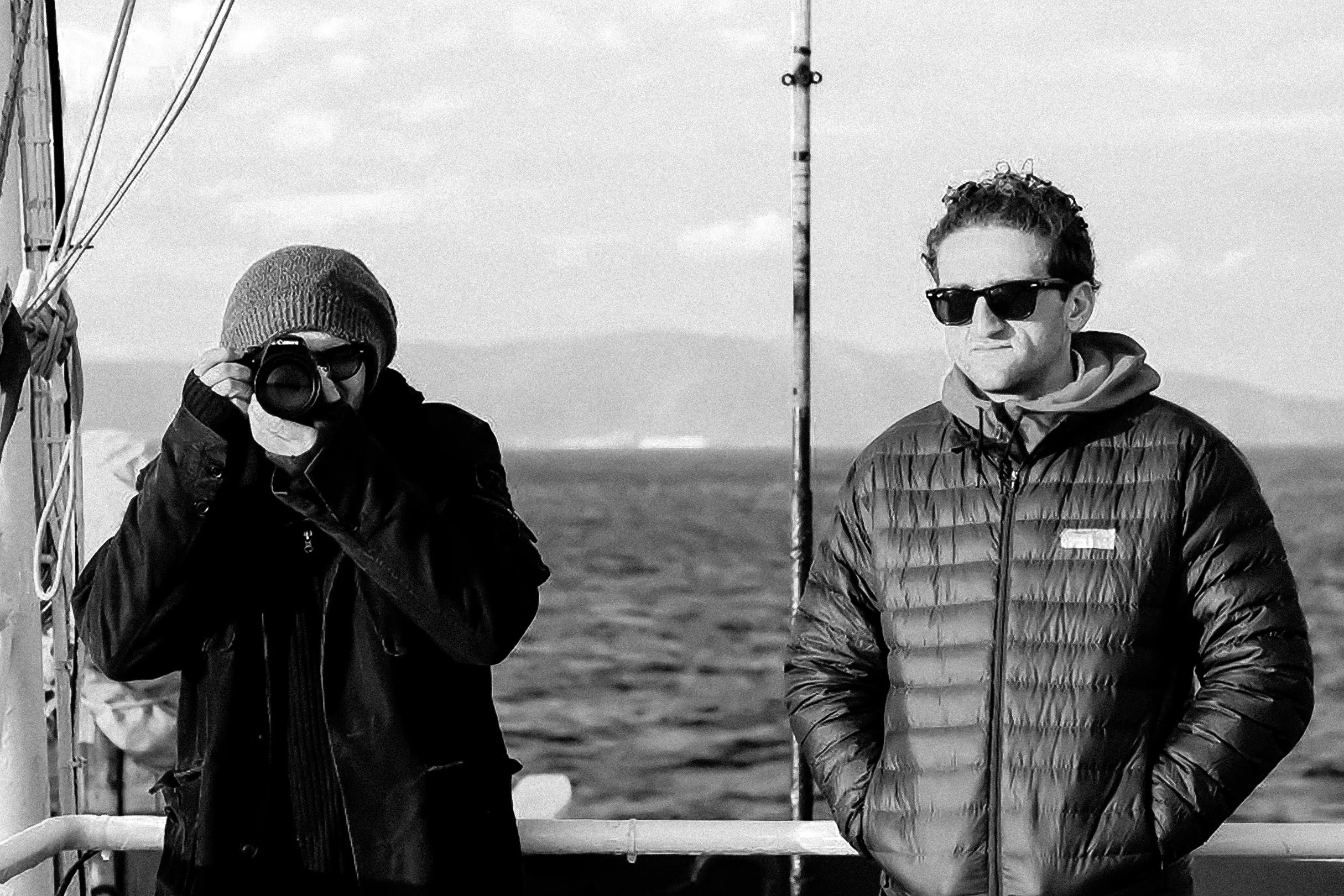 Two people stand outdoors on a boat, one holding a camera and the other standing beside a pole, with the ocean and cloudy sky in the background.