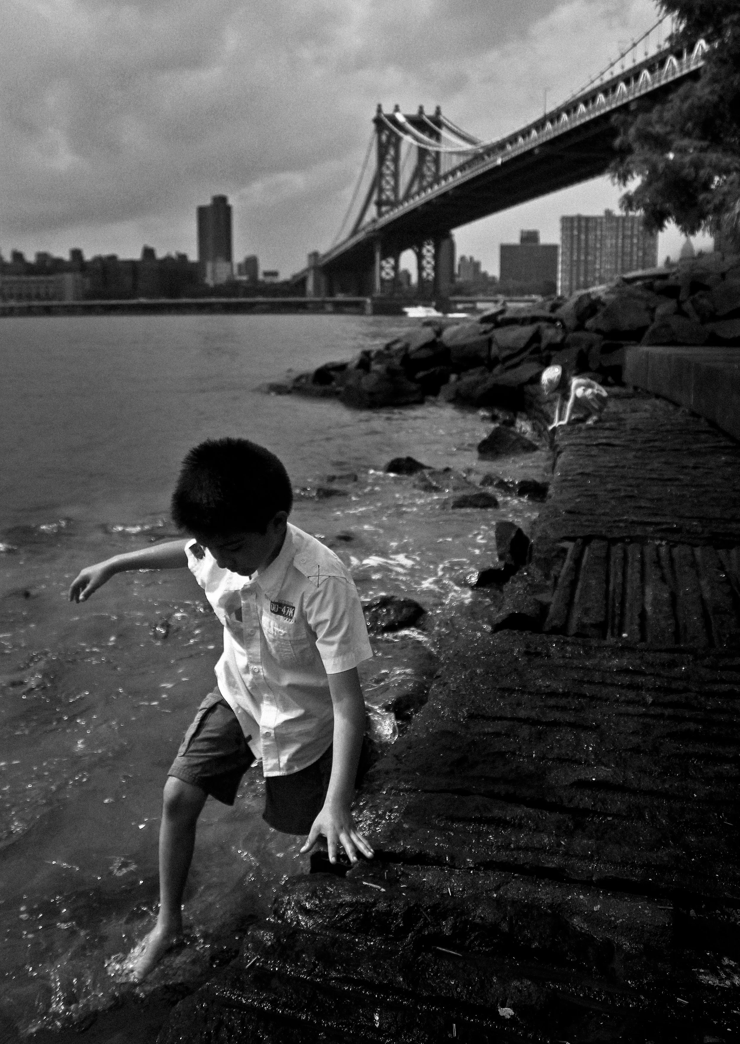 A black and white photo of a boy wading in water near a rocky shoreline with a city skyline and a bridge in the background.