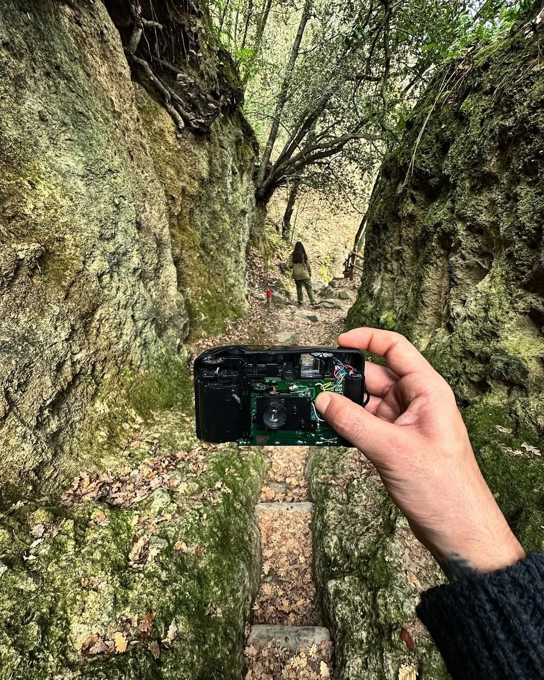 A person taking a photograph with a camera in a narrow, moss-covered rocky hiking trail surrounded by trees.