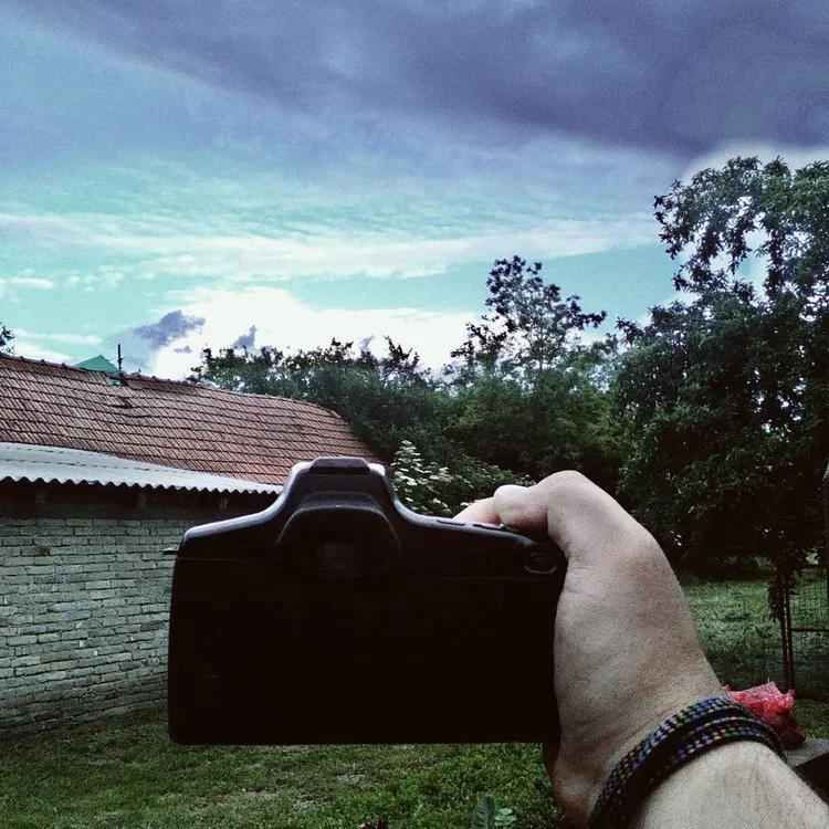 Person holding a camera in front of house and trees with a cloudy sky in the background