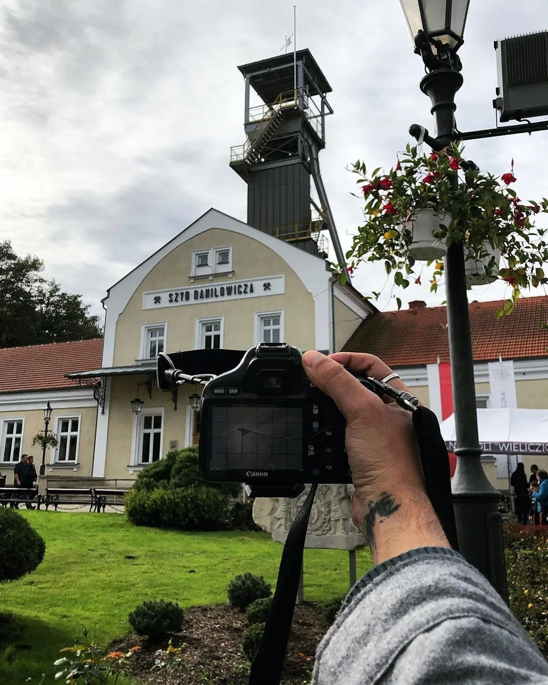 Person holding a camera taking a photo of a historic building with a tower and the sign 'Szyd Damilowicza' on the facade, with a streetlamp, bushes, and people in the background.