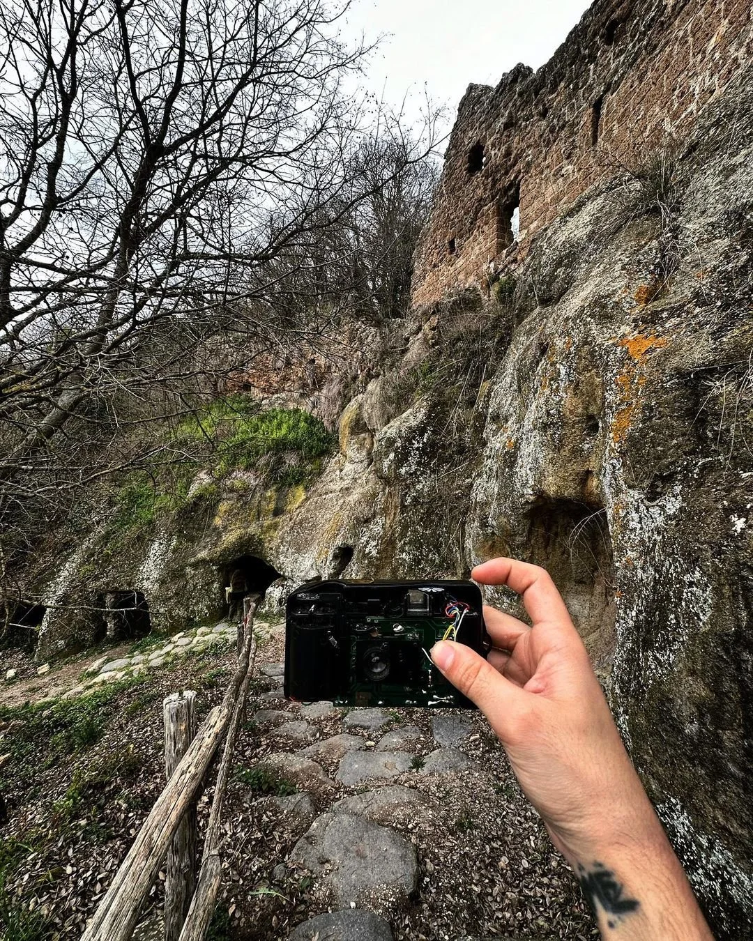 Person taking a photograph of a rocky hillside with a camera, with leafless trees and an old stone structure at the top of the hill.