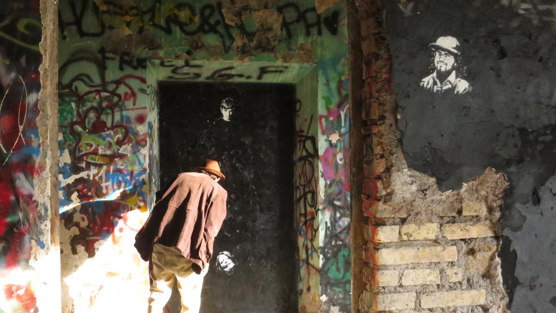 A person with a brown hat and oversized coat exploring an abandoned, graffiti-covered underground tunnel with black and multicolored walls.