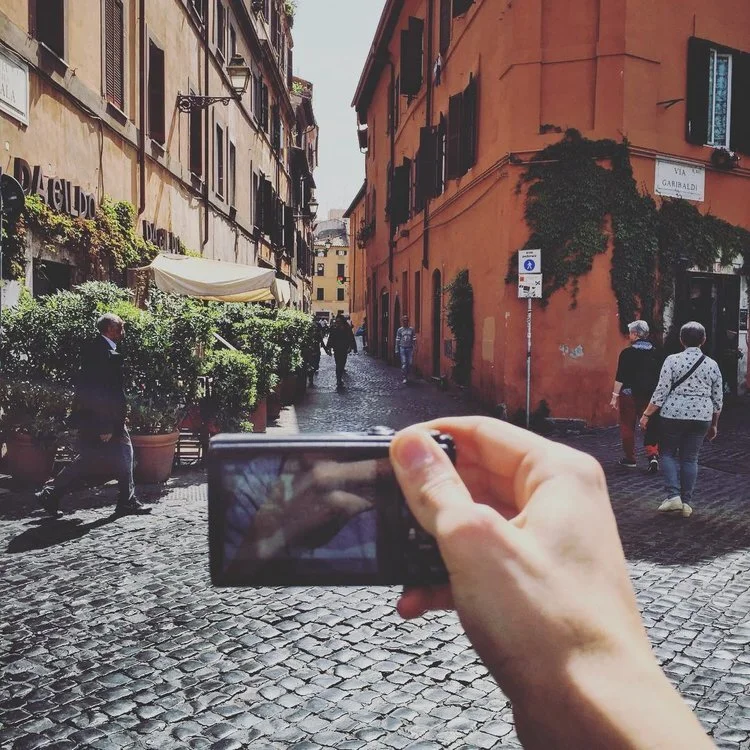 A person is taking a photo of a cobblestone street scene with a digital camera. The street is lined with colorful old buildings, some with green plants and awnings. Several people are walking along the street, and there are potted plants outside one 