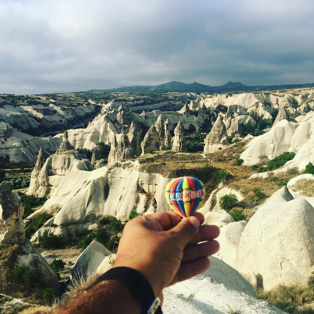 Person holding a small colorful hot air balloon figurine in front of Cappadocia's unique rock formations and fairy chimneys in Turkey.