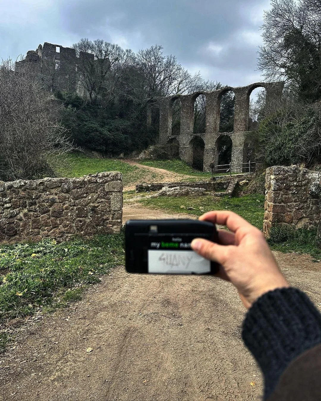 Person taking a photo of ancient stone ruins and an aqueduct on a dirt path on a cloudy day.