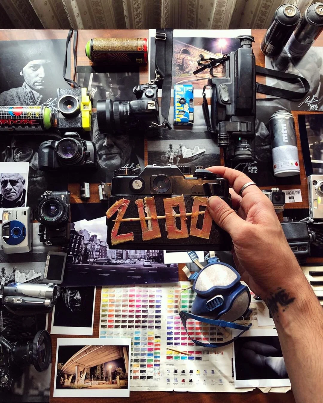 A cluttered photography workspace with cameras, film, photos, and art supplies. A hand holds a camera with 'ZUBO' spelled out on it.