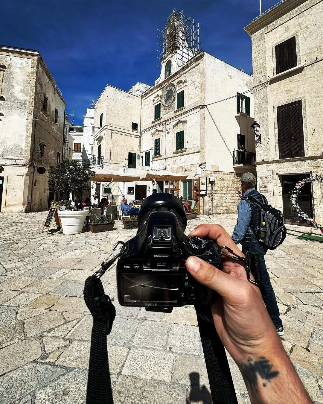 A person holds a camera while taking a photo of a European town square with outdoor cafe seating, a white historic building with scaffolding, and people sitting at tables on a sunny day.