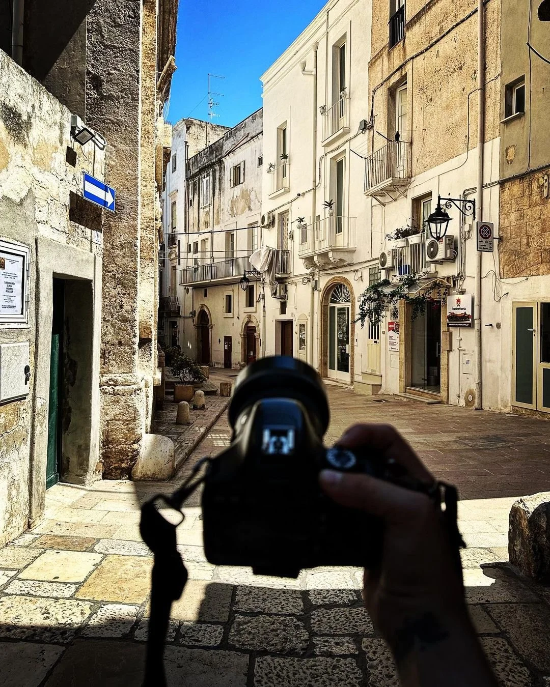 A person holding a camera, taking a photo of a narrow, historic street in a European town with old stone and stucco buildings, balconies, air conditioning units, potted plants, and a clear blue sky.
