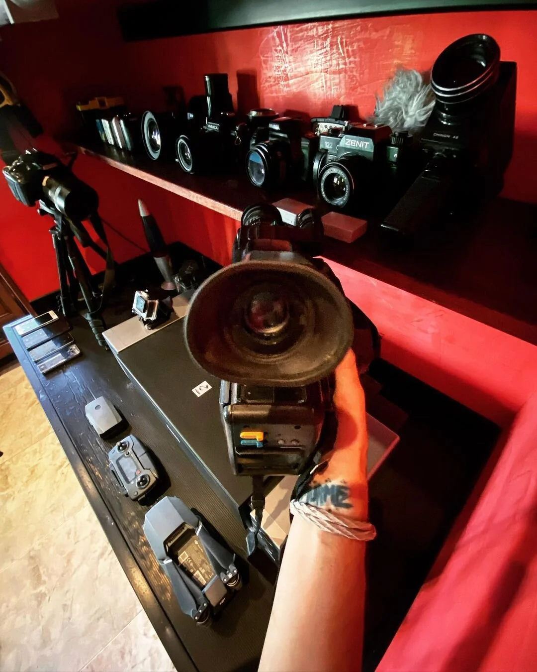 Camera equipment including various cameras on a shelf and a person holding a camera, with drone and remote controls on a table, set against a red wall inside a room.