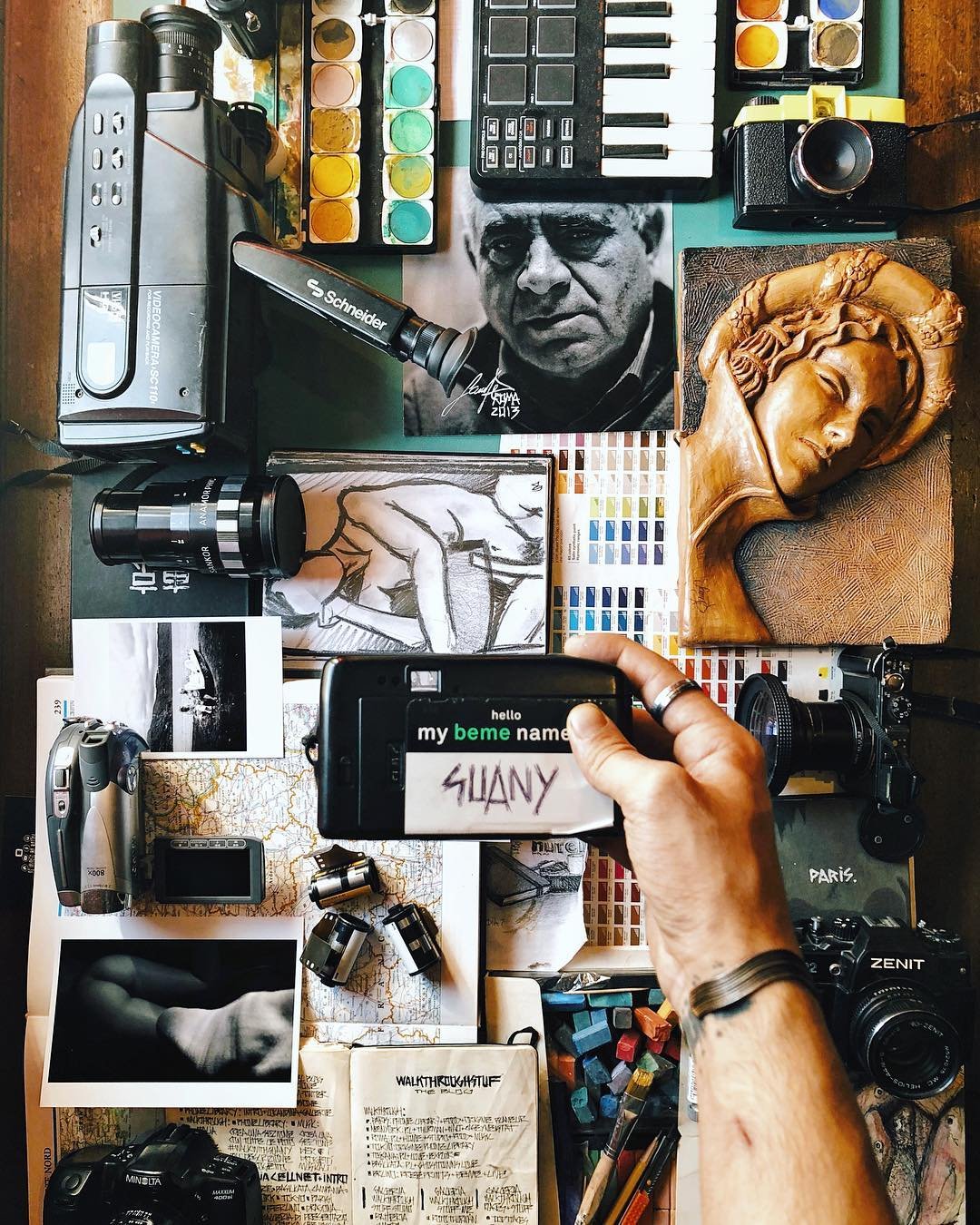 A top-down view of a cluttered desk with photography and art supplies, including cameras, paint palettes, sketches, a wooden sculpture, a framed black and white photo, a printed map, and a person's hand holding a camera with a sign displaying 'my name' as 'SuanY'.
