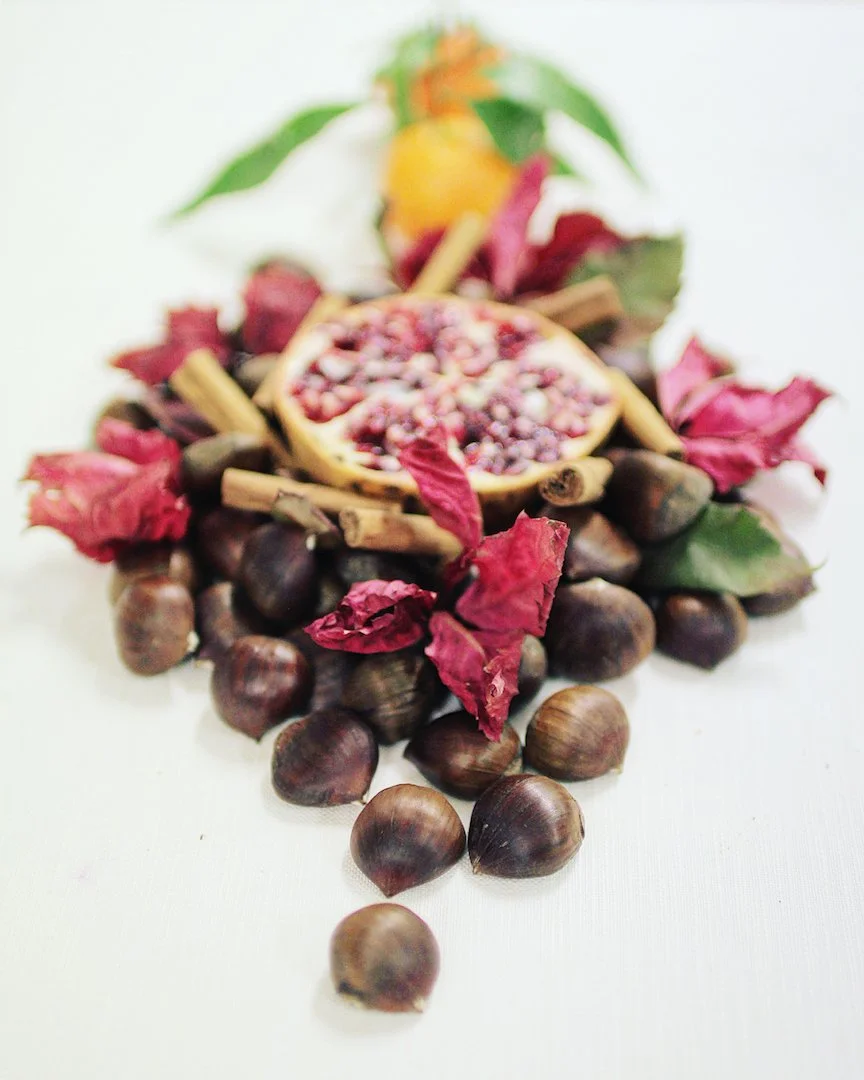 A halved pomegranate surrounded by hazelnuts, dried rose petals, cinnamon sticks, and a sprig of green leaves