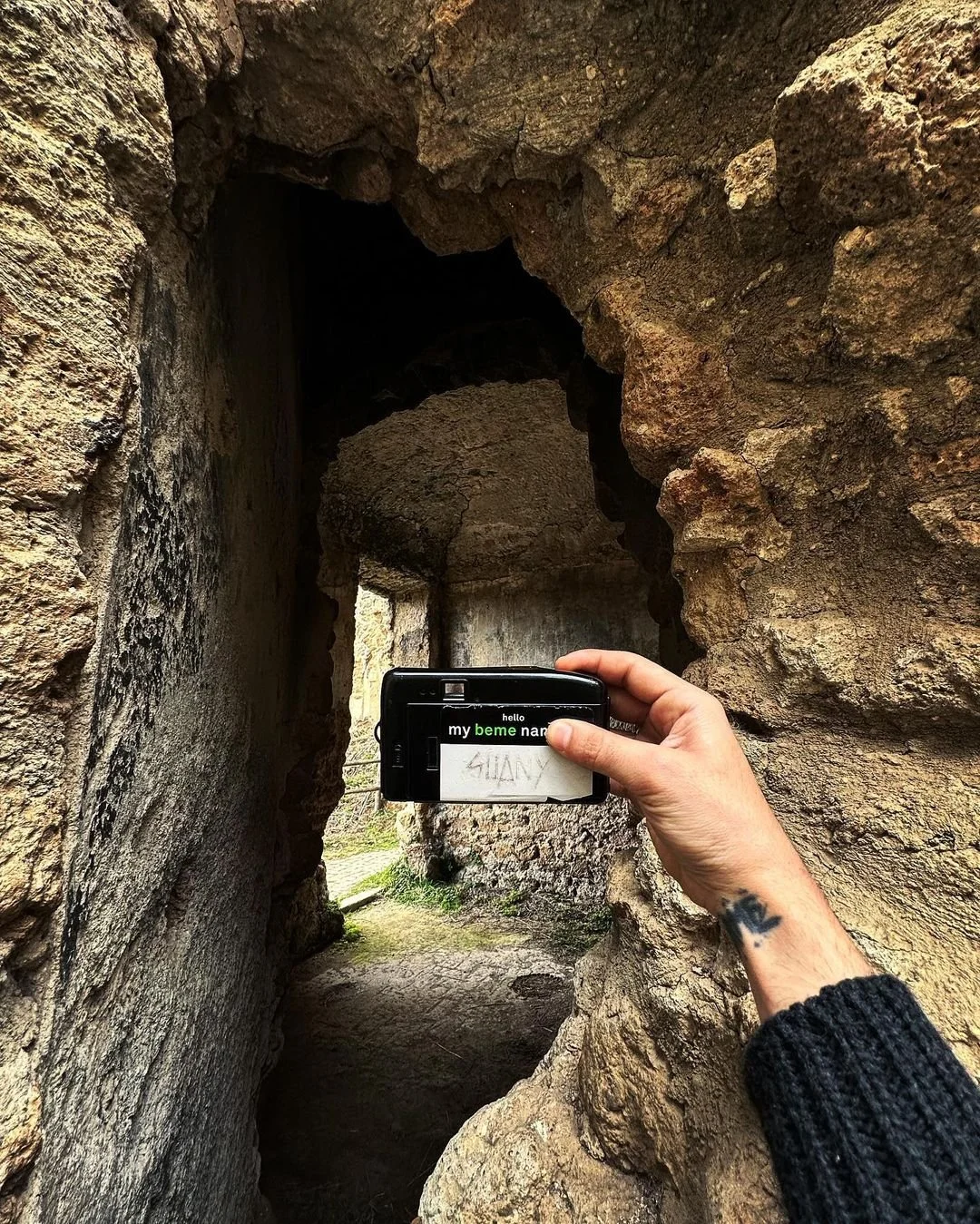 Person taking a photo with a digital camera of a rocky, ancient-looking stone archway in a narrow corridor.