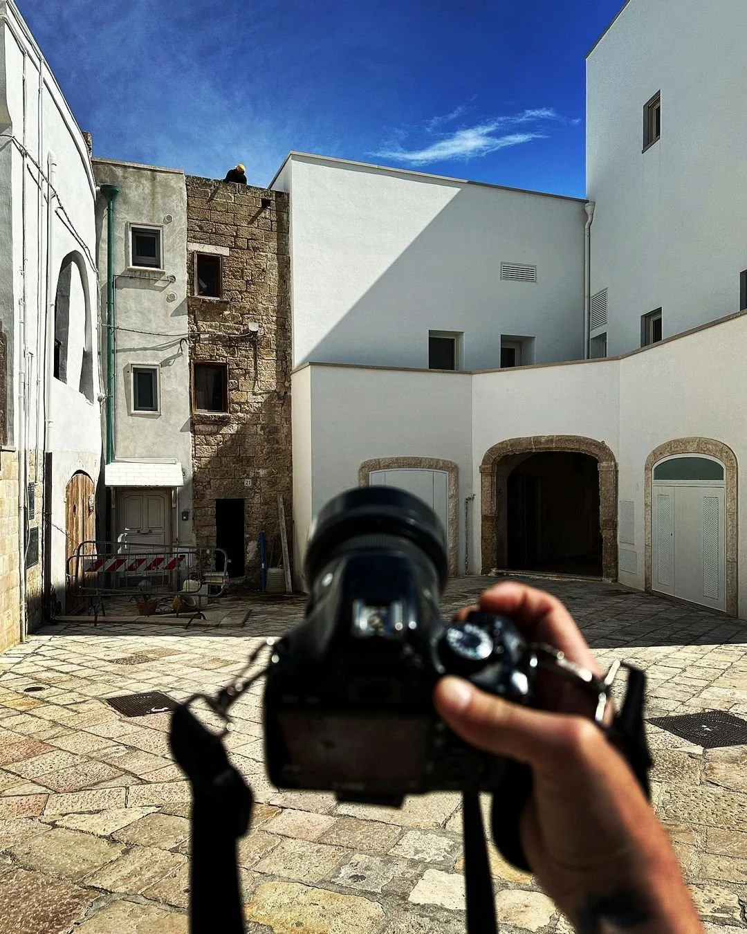 A person holding a camera, taking a photo of a small courtyard with stone pavement, white and stone buildings, and a blue sky with some clouds.