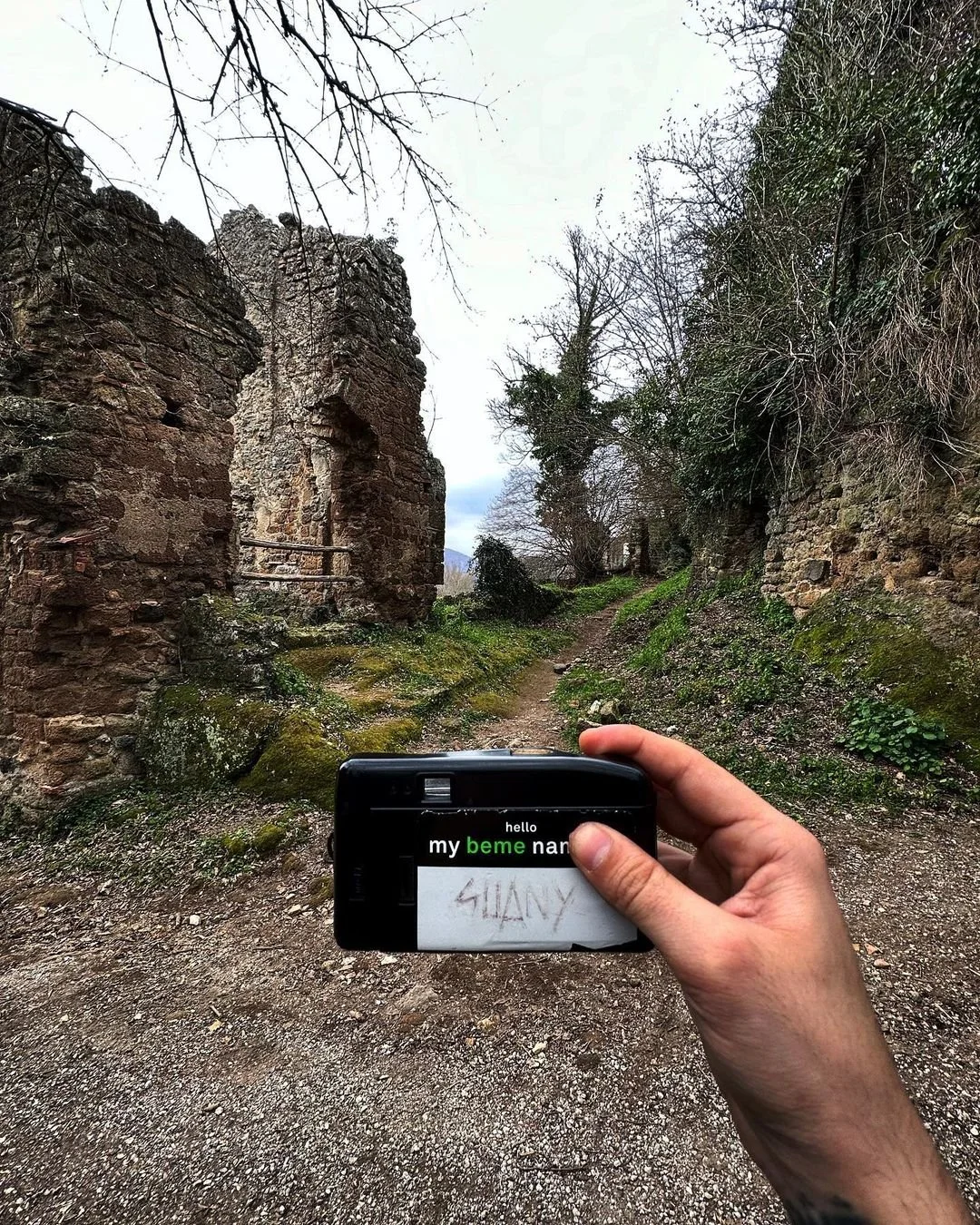 A camera held by a person captures an image of an old, ruined stone building on a dirt path, with trees and greenery surrounding the scene.