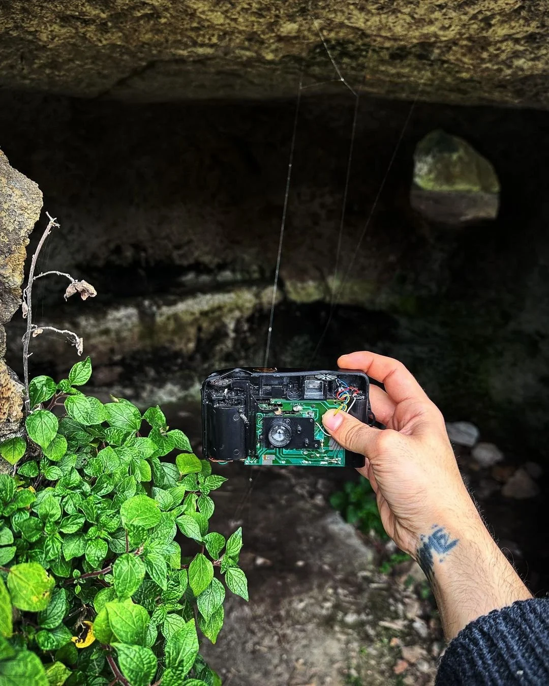 A person holding a broken camera with exposed circuit board near a natural cave or tunnel with greenery in the foreground.