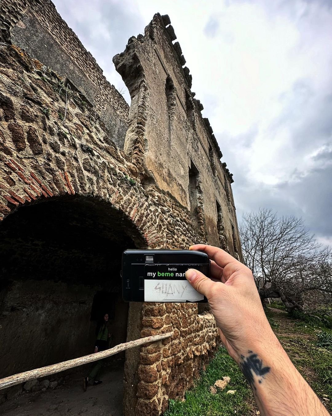 Person taking a photo of an old stone and brick building with a smartphone. The building has an archway entrance and worn walls, with a cloudy sky above and a person visible in the background under the arch.