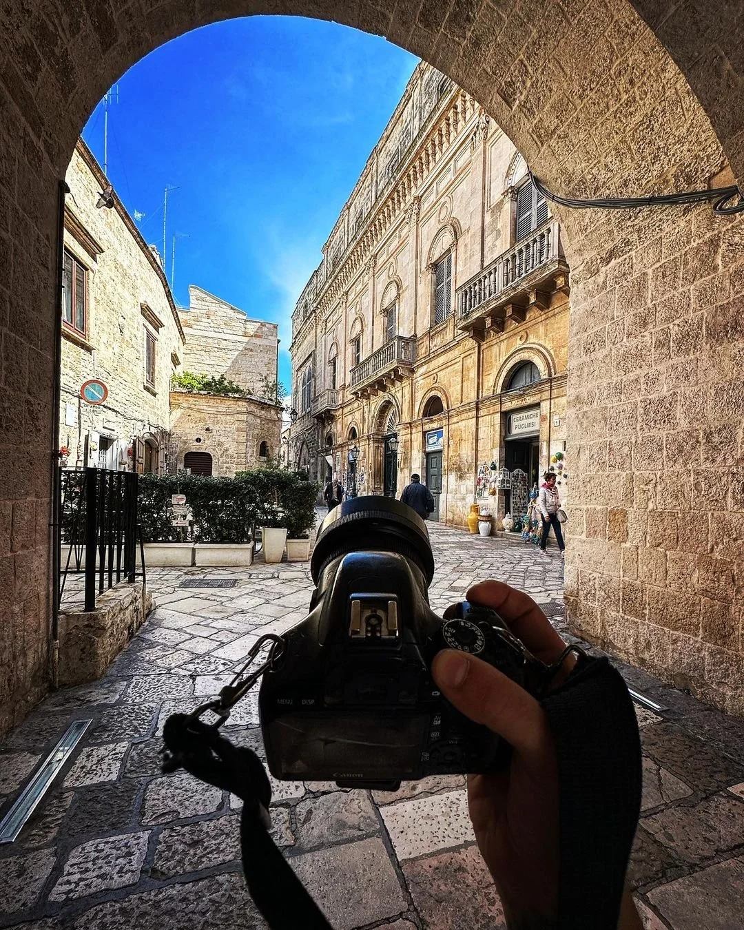 A street scene viewed through an archway with a person taking a photo of the historic buildings and pedestrians outside under a bright blue sky.
