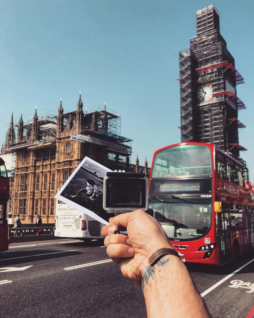 A person holding a black and white photograph of a woman on a beach in front of Big Ben in London, with red double-decker buses and legal building in the background.