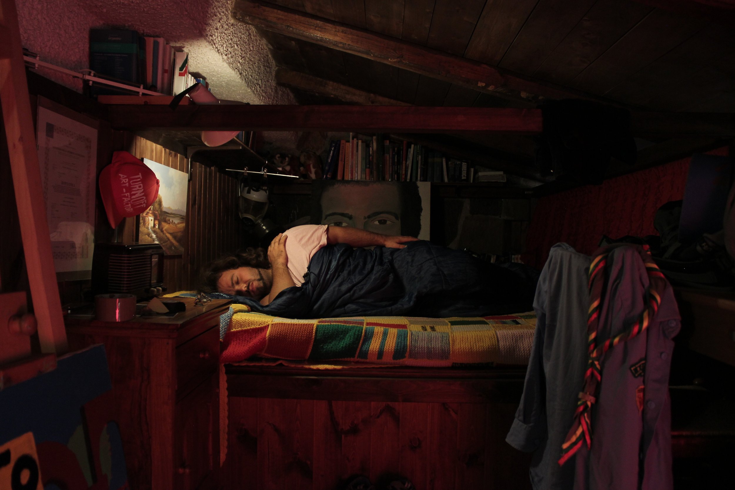 A person with curly hair and a beard is lying down on a bed, resting with their head on a pillow, inside a cozy, wooden-paneled room. The room contains books on a shelf, a painting of a landscape with a house, and various personal items around.